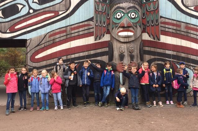 Gruppe von Kindern vor einer kunstvoll bemalten Totempfahlwand im Freien, l&auml;chelnd und teils posierend.