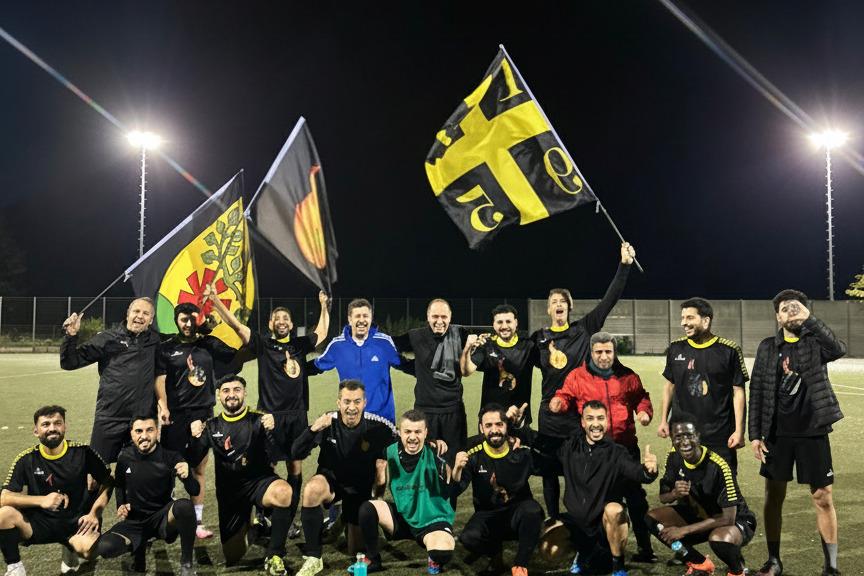 Gruppe von Fu&szlig;ballspielern in schwarzen Trikots posiert feiernd auf einem Spielfeld, w&auml;hrend sie Fahnen halten.