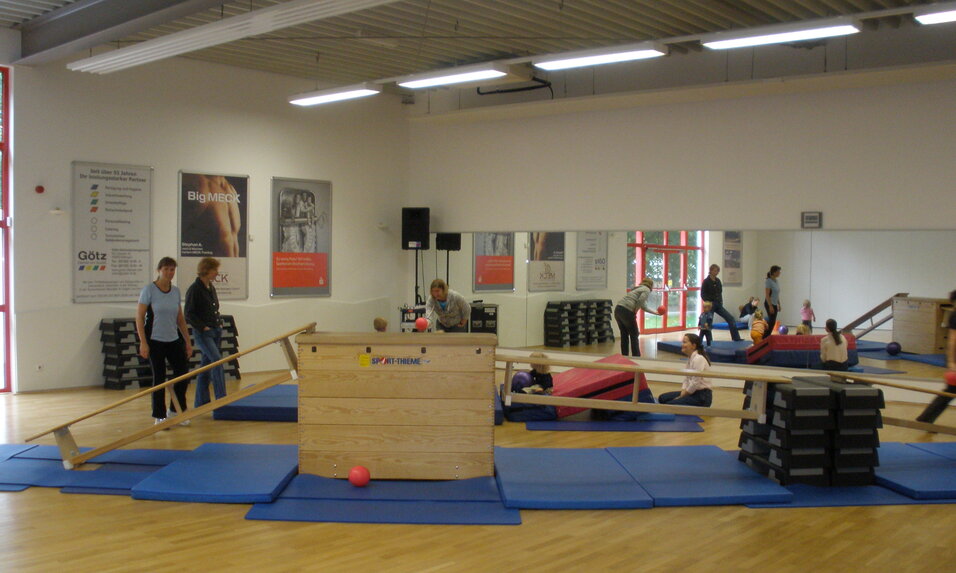 Sportliche &Uuml;bungseinheit in einem Trainingsraum mit Ger&auml;ten, Matten und mehreren Personen im Hintergrund.