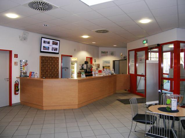 Empfangsbereich mit einer Holztheke, Informationen und Sitzgelegenheiten, gro&szlig;en Fenstern und roten Akzenten.