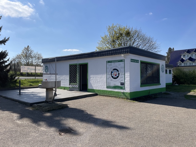 Ein schlichtes Geb&auml;ude mit gr&uuml;ner Fassade, Fenstern und einem dartspielenden Plakat an einer Freifl&auml;che bei sch&ouml;nem Wetter.