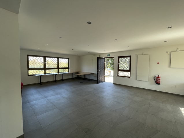 Heller, leerer Raum mit Fliesenboden, zwei Fenstern und einem Eingangst&uuml;r. Ein Tisch steht im Hintergrund.