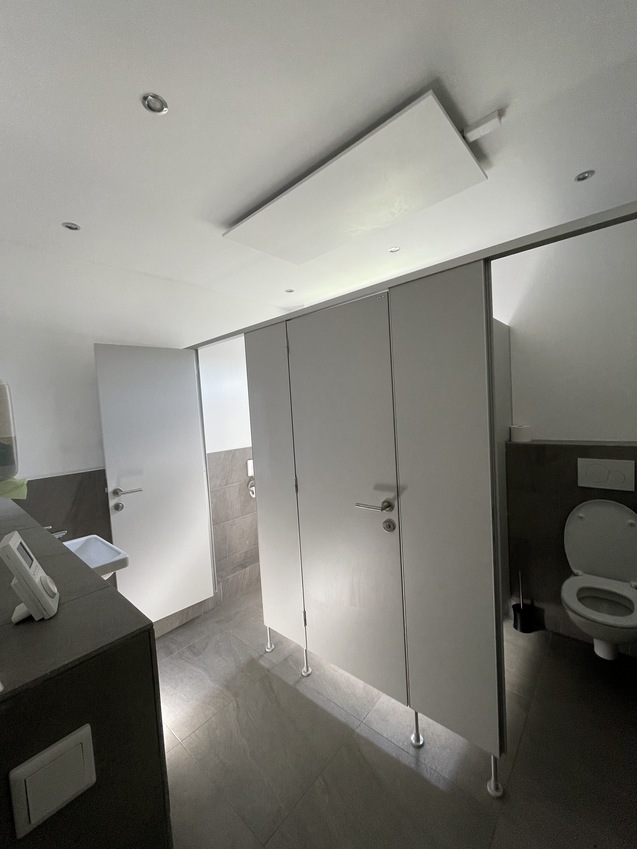 Blick in ein modernes, beleuchtetes Badezimmer mit Trennw&auml;nden, Waschbecken und einer Toilette.