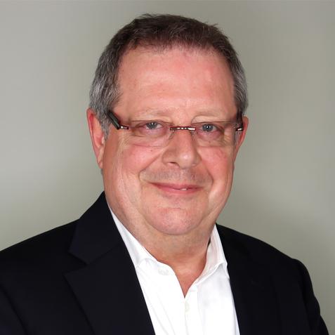 Portr&auml;t eines l&auml;chelnden Mannes mittleren Alters mit Brille, in Anzugjacke und wei&szlig;em Hemd vor einem grauen Hintergrund.