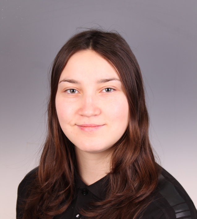 Portr&auml;t einer Person mit langen, braunen Haaren, die ein schwarzes T-Shirt mit einem Aufdruck tr&auml;gt. Hintergrund grau.