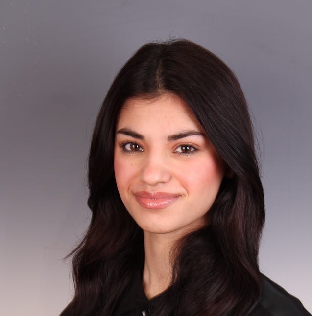 Portr&auml;t einer jungen Frau mit langen, dunklen Haaren, die in einem schwarzen Oberteil vor einem neutralen Hintergrund l&auml;chelt.