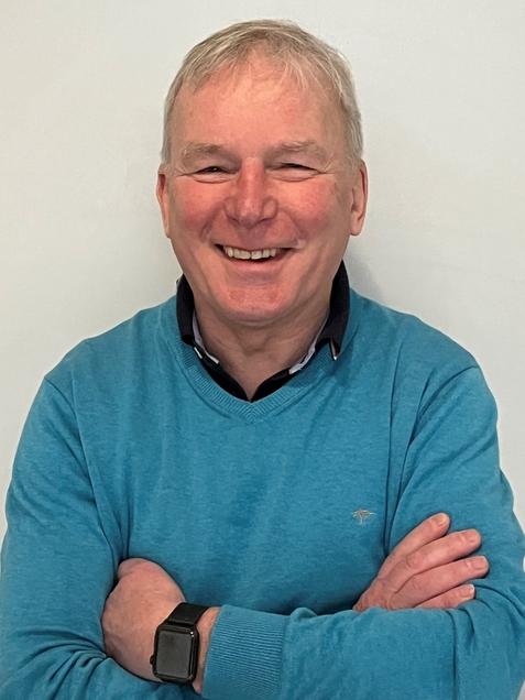 Lächelnder Mann mit kurzen Haaren, in blauer Pullover, steht mit verschränkten Armen vor einer hellen Wand.