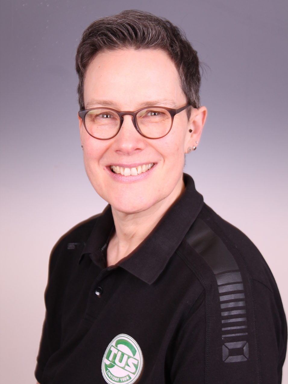 Portrait einer l&auml;chelnden Frau mit kurzen Haaren und Brille, im schwarzen Poloshirt mit TUS-Logo.