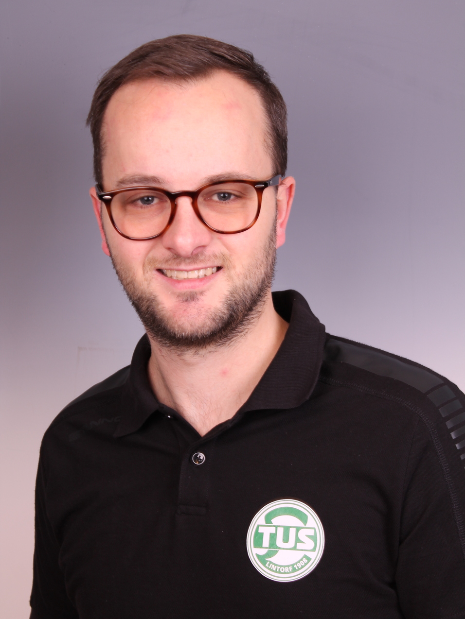 Portrait eines Mannes mit Brille, der ein schwarzes Polo-Shirt mit Logo tr&auml;gt, vor grauem Hintergrund.