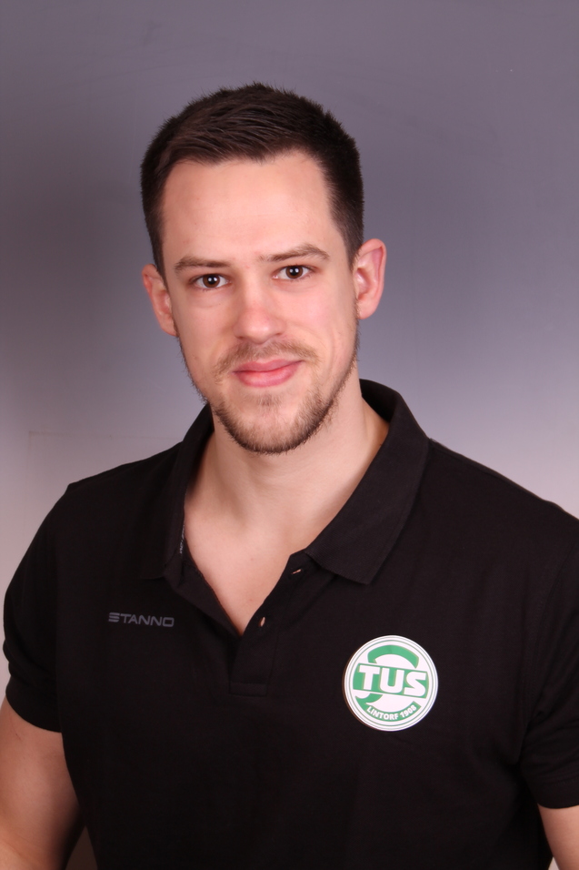 Portr&auml;t eines l&auml;chelnden Mannes in schwarzem Polo-Shirt mit &bdquo;TUS&ldquo;-Logo auf grauem Hintergrund.