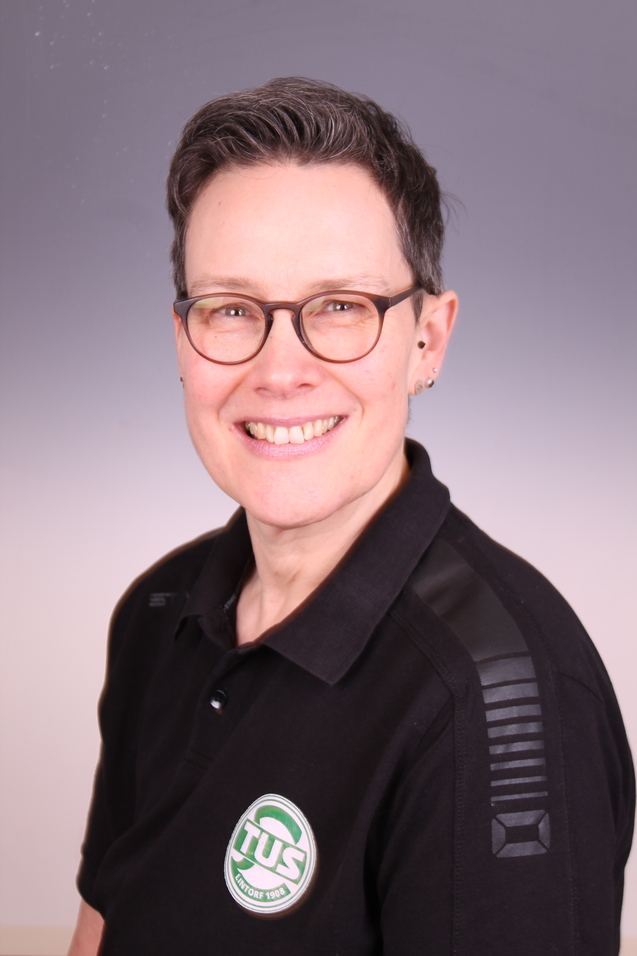 Portrait einer l&auml;chelnden Frau mit kurzen Haaren und Brille, im schwarzen Poloshirt mit TUS-Logo.