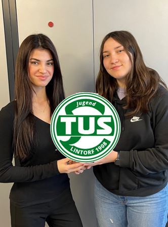 Zwei junge Frauen geben sich die Hand vor grauem Hintergrund, mit dem Vereinslogo von TuS Lintorf 1908 sichtbar.