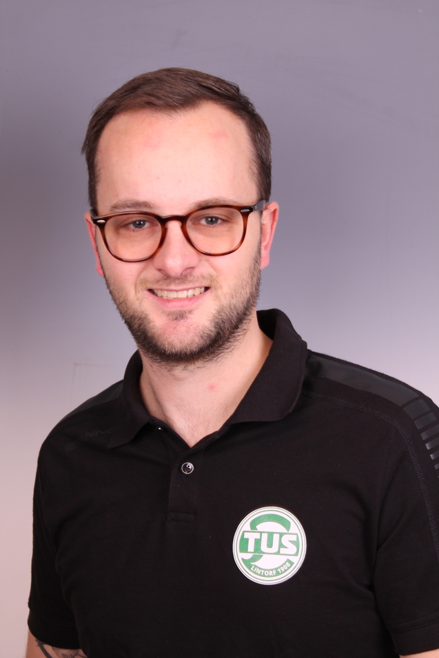 Portrait eines Mannes mit Brille, der ein schwarzes Polo-Shirt mit Logo tr&auml;gt, vor grauem Hintergrund.