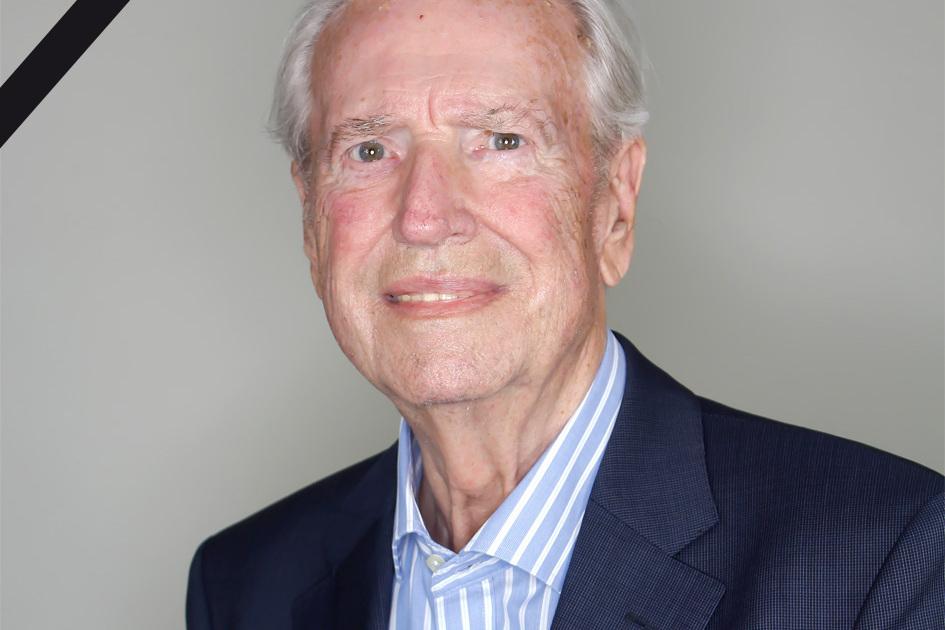 Portr&auml;t eines &auml;lteren Mannes mit grauen Haaren, in einem Anzug, l&auml;chelt vor einem neutralen Hintergrund mit Trauerrand.