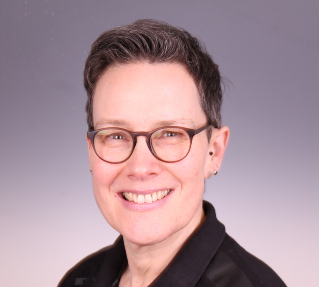 Portrait einer l&auml;chelnden Frau mit kurzen Haaren und Brille, im schwarzen Poloshirt mit TUS-Logo.
