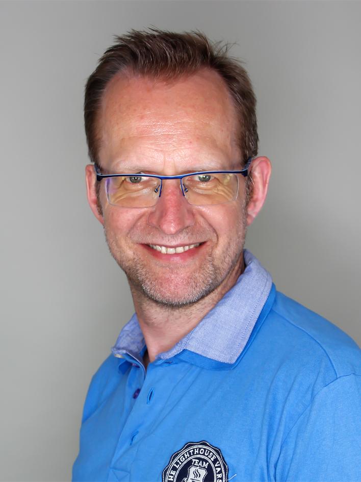 Lächelnder Mann mit Brille und kurzem Bart trägt ein hellblaues Poloshirt, Hintergrund grau.