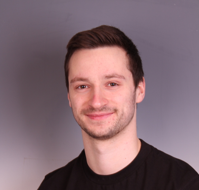 Portr&auml;t eines l&auml;chelnden Mannes mit kurzem Haar, der ein schwarzes T-Shirt mit einem Logo tr&auml;gt. Hintergrund in Grau.