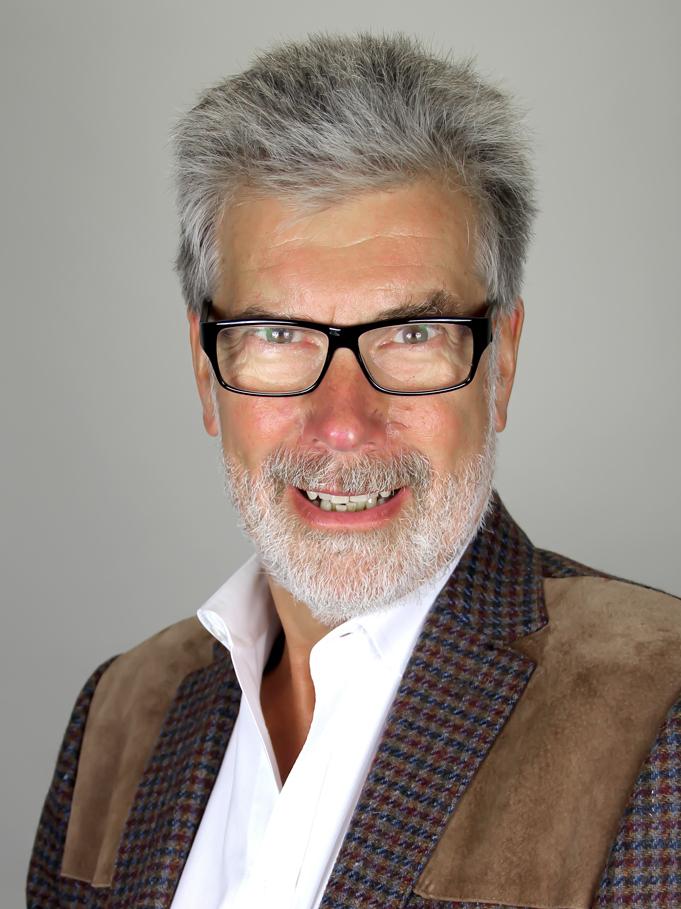 Helle Portraitaufnahme eines lächelnden Mannes mit Brille, grauem Haar und kariertem Sakko vor neutralem Hintergrund.