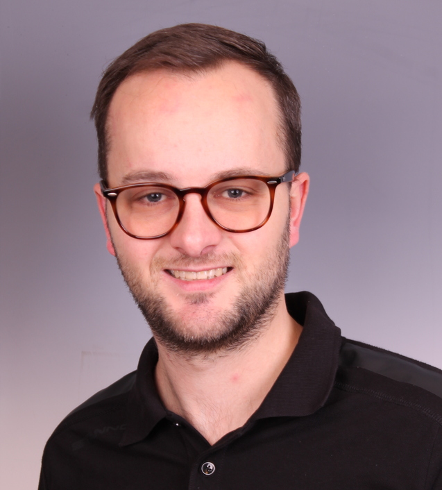 Portrait eines Mannes mit Brille, der ein schwarzes Polo-Shirt mit Logo tr&auml;gt, vor grauem Hintergrund.