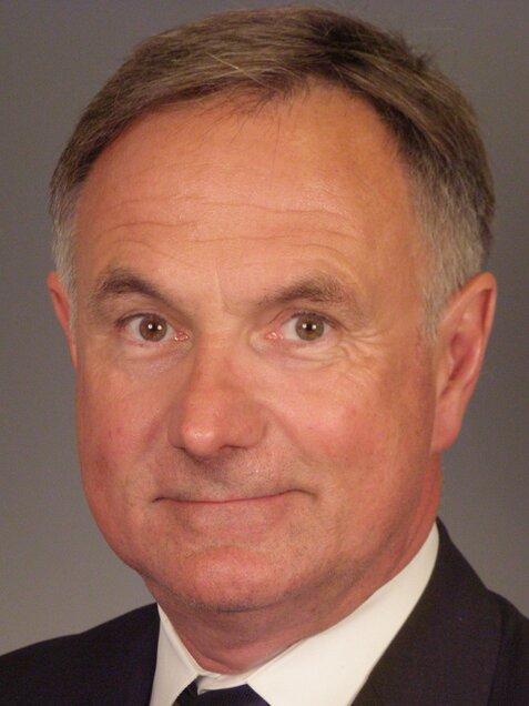 Ein Mann mit grauen Haaren und Anzug schaut in die Kamera, Hintergrund in neutralem Grau.