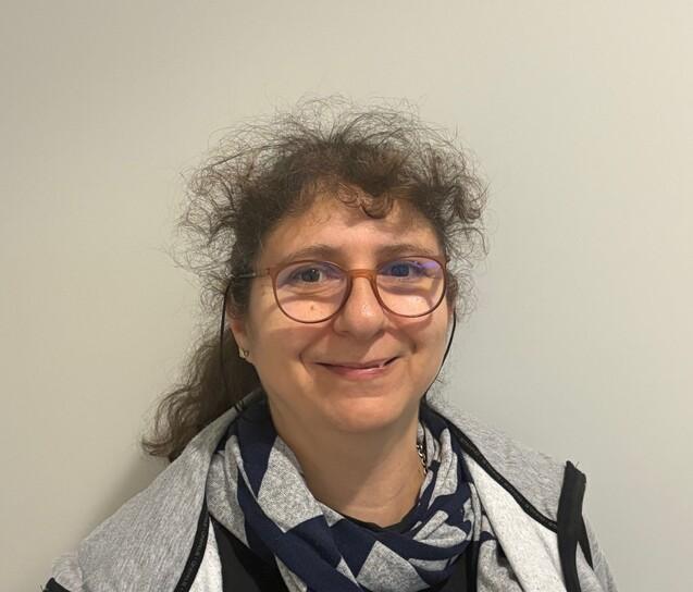 Frau mit lockigem Haar und Brille trägt einen grauen Hoodie und ein gestreiftes Halstuch vor neutralem Hintergrund.