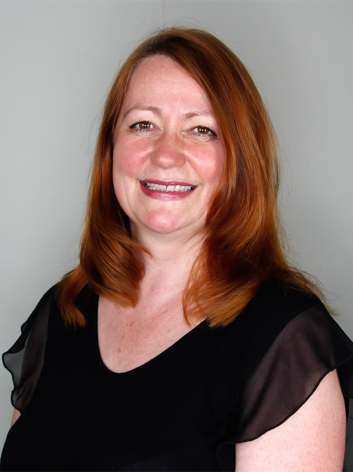 Porträt einer lächelnden Frau mit rotbraunem Haar, die ein schwarzes Oberteil trägt, vor grauem Hintergrund.