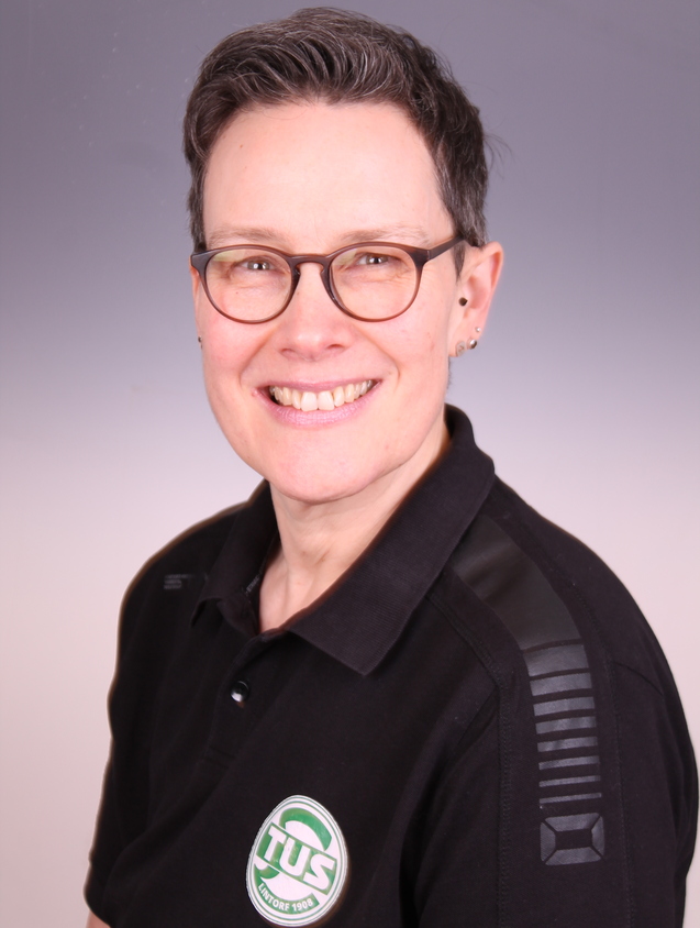 Portrait einer l&auml;chelnden Frau mit kurzen Haaren und Brille, im schwarzen Poloshirt mit TUS-Logo.