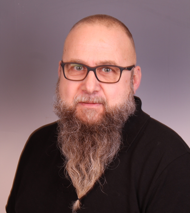 Mann mit Brille und langen Bart, tr&auml;gt schwarzes Polohemd, posiert vor neutralem Hintergrund.