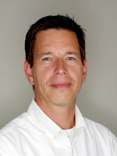 Porträt eines Mannes mit kurzem, braunem Haar in einem weißen Hemd vor einem grauen Hintergrund.